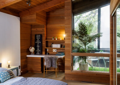 A cozy, rustic modern bedroom featuring extensive wood paneling and exposed beams on the ceiling and walls. A large angled window overlooks an outdoor patio with a palm plant inside. The room includes a bed with a grey comforter and a built-in desk area with shelves, a vintage typewriter, and a furry chair.