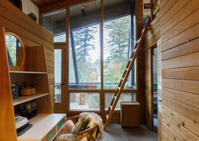 Rustic modern home office with wood paneling, vaulted ceiling, large windows overlooking trees, a desk, faux fur chair, and a wooden ladder to a loft.