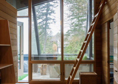 Modern wood-paneled room with large windows, a built-in desk, a wooden ladder, and a view of a forest.