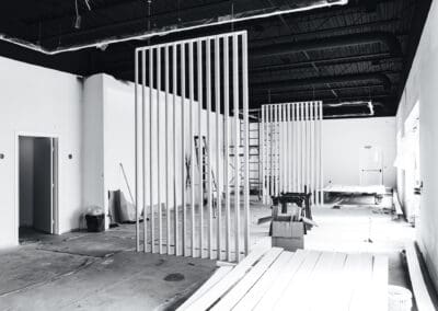 Black and white photo of an interior commercial space under renovation with vertical slat walls, exposed industrial ceiling, and construction materials.