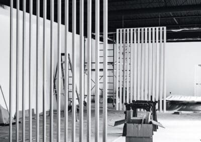 Black and white photo of an unfinished modern interior construction site featuring vertical slat partitions, exposed industrial ceiling, scaffolding, and building materials.