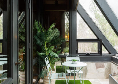 Modern sunroom patio with mint green bistro set, artificial turf rug, and potted palm plants under large glass windows.