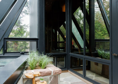 Modern outdoor patio with a unique wooden plant stand, flagstone paving, and large angular glass windows overlooking a wooded area.