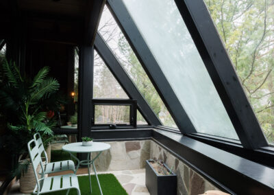 Modern sunroom or enclosed patio with large sloped glass windows, featuring a light green metal bistro set on a small turf rug, natural stone flooring, and rustic wood accents amidst lush greenery.