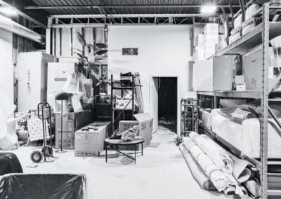 Black and white warehouse interior with furniture storage, stacked boxes, shelving units, and wrapped items.