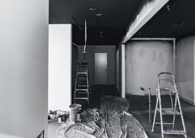 Black and white photo of an unfinished room under renovation, with paint cans, drop cloths, and two ladders. The walls are partially prepped, and the ceiling is dark.