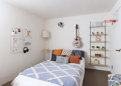 Modern white bedroom with a bed featuring blue and white patterned bedding, an acoustic guitar, wall shelves with plants and books, and a mini basketball hoop.
