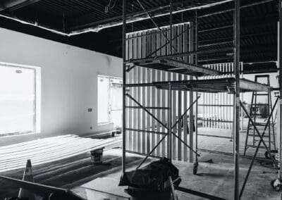 Black and white interior photo of an active construction site with metal scaffolding, unfinished walls, covered windows, and wooden trim on the floor.