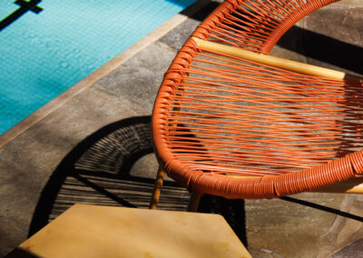 Vibrant orange woven patio chair and a hexagonal yellow side table next to a sparkling blue swimming pool, featuring strong sunlight and shadows on the stone deck.