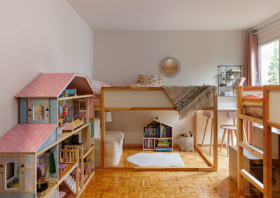 A child's bright and organized bedroom featuring a large multi-story dollhouse, a wooden loft bed, a desk area, and a cozy reading nook with a house-shaped bookshelf and a white rug.