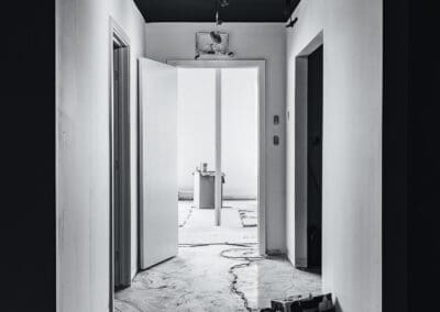 Black and white photo of a hallway under renovation, featuring a dark ceiling, white walls, open doors, and painting supplies on the floor.