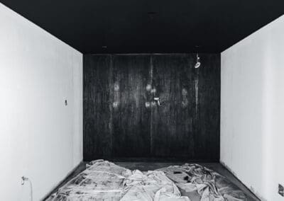 Monochrome image of an empty room under renovation with white walls, a black ceiling, and dark wood paneled back wall, with drop cloths on the floor.