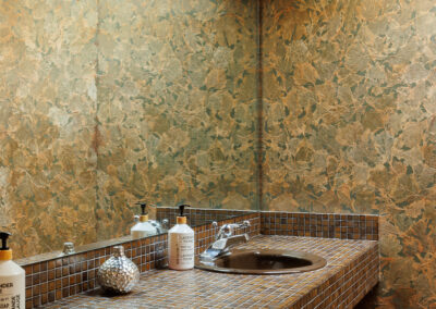 Unique bathroom with double dark sinks, a brown mosaic tile countertop, and distinctive green and golden leaf-patterned wallpaper under a skylight.