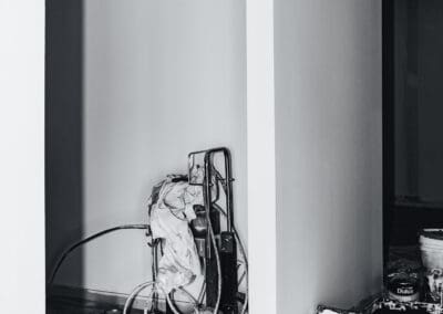 Black and white image showing an airless paint sprayer, paint roller, and Dulux paint can on drop cloths in a room undergoing interior painting or renovation.