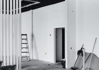 Black and white photo of an unfinished commercial interior under renovation, featuring a slat wall partition, an A-frame ladder, exposed ceiling ducts, and construction tools on a concrete floor.