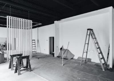 Black and white image of an unfinished commercial interior under renovation, featuring white walls, a dark ceiling, a modern wooden slat room divider, two ladders, and two Stanley saw horses on a concrete floor.