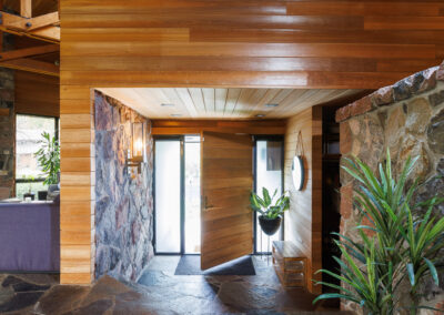 Rustic modern home interior with exposed wood beams, stone accent walls, flagstone flooring, and an open wooden front door. Natural light enters through skylights.