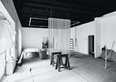 Black and white image of a modern commercial interior under construction, featuring white walls, an exposed black ceiling with ductwork, and a slatted room divider with saw horses and tools on the protective floor.