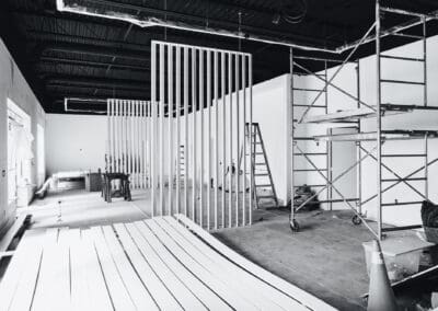 Black and white interior photo of a commercial space under renovation, featuring scaffolding, a ladder, partially installed vertical slat wall panels, and white trim pieces on the floor. Exposed industrial-style ceiling with ductwork is visible.