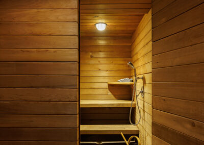 Interior view of a modern wooden sauna room featuring horizontal wood paneling, tiered benches, a wall-mounted shower, and a stone floor. A black sauna bucket with a yellow handle and ladle is visible near the entrance.