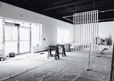 Black and white photo of an interior commercial construction site with exposed ceiling, large plastic-covered windows, saw horses, and a modern slat room divider.