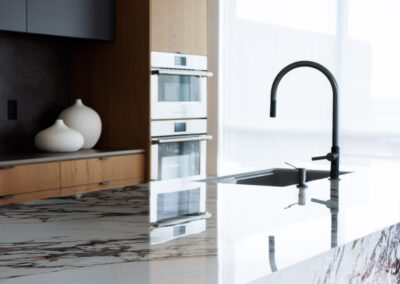 Modern kitchen island with white and reddish-brown veined marble countertop, sleek black faucet, built-in ovens, and natural wood cabinetry.