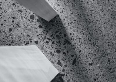 Monochromatic image of a grey terrazzo surface with dark aggregate, featuring diagonal white architectural elements casting sharp shadows.