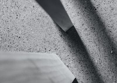 Monochromatic image of three light-colored rectangular beams casting sharp, dark geometric shadows on a speckled grey terrazzo or concrete floor.