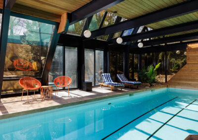 Modern indoor swimming pool with glass enclosure, wooden ceiling, orange wicker chairs, and striped lounge chairs overlooking lush greenery.