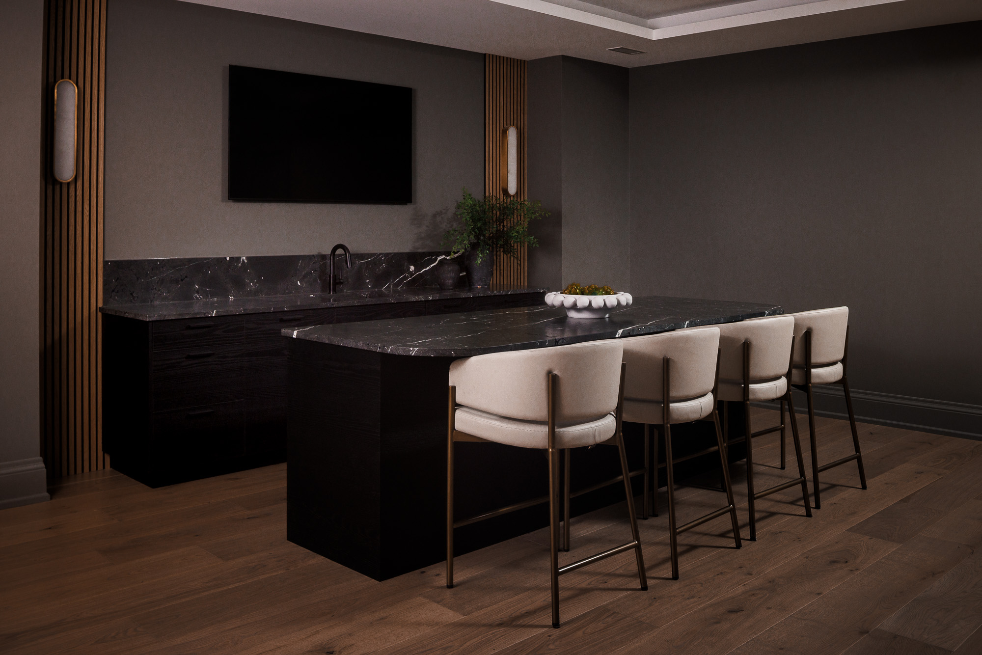 2A4A8084 A bright, modern kitchen featuring a dark green countertop on a ribbed wooden island with black bar stools, a brass gooseneck faucet above a sink by a large window, light cream cabinetry, and a range cooker set against a veined marble backsplash under a custom hood.