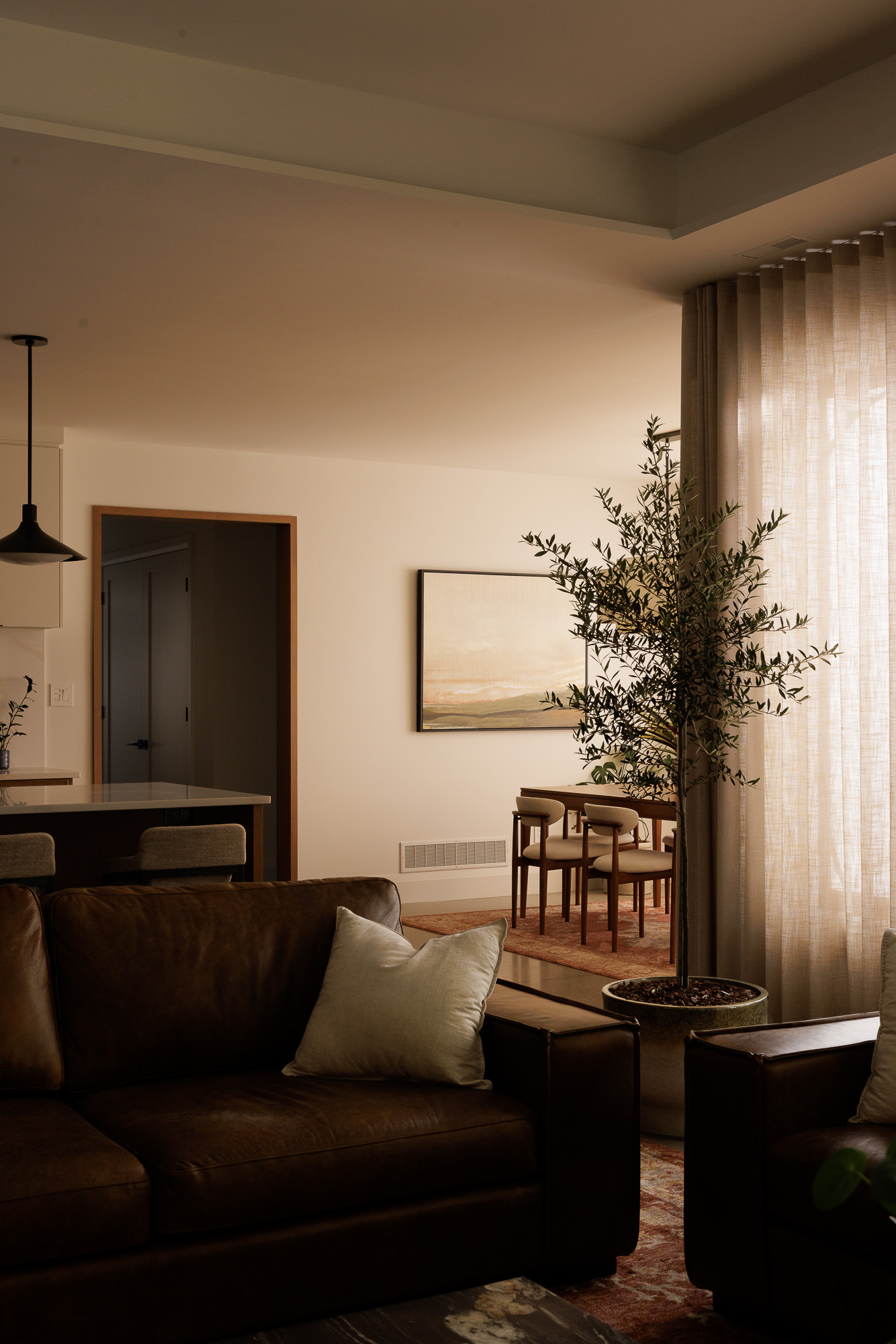Modern dining room with a long wooden table, potted plant, cream fabric pendant light, sheer beige ripple fold curtains, and an antique-style area rug.