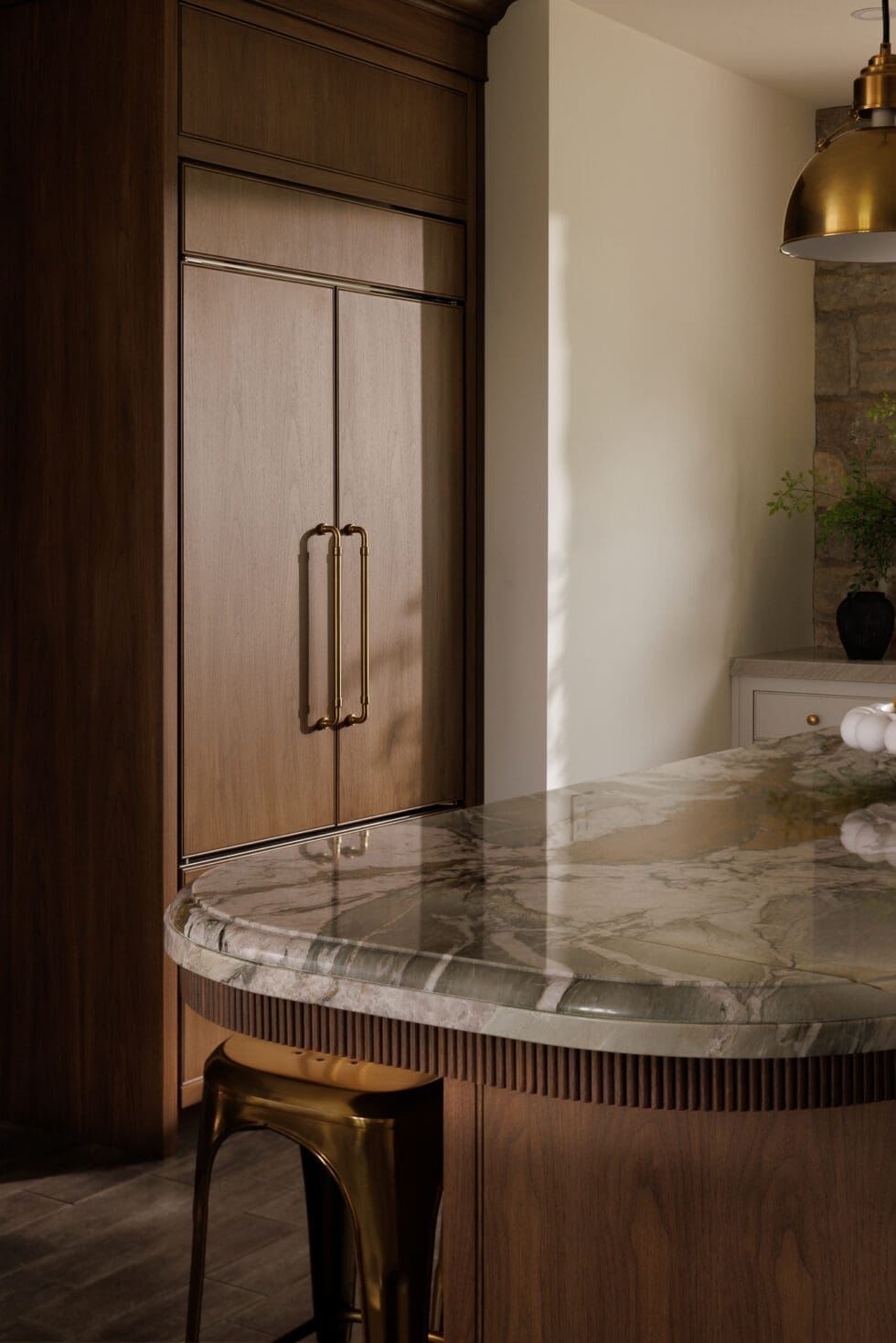 Elegant dark wood paneled built-in refrigerator with brass handles next to a kitchen island with veined green marble countertop.