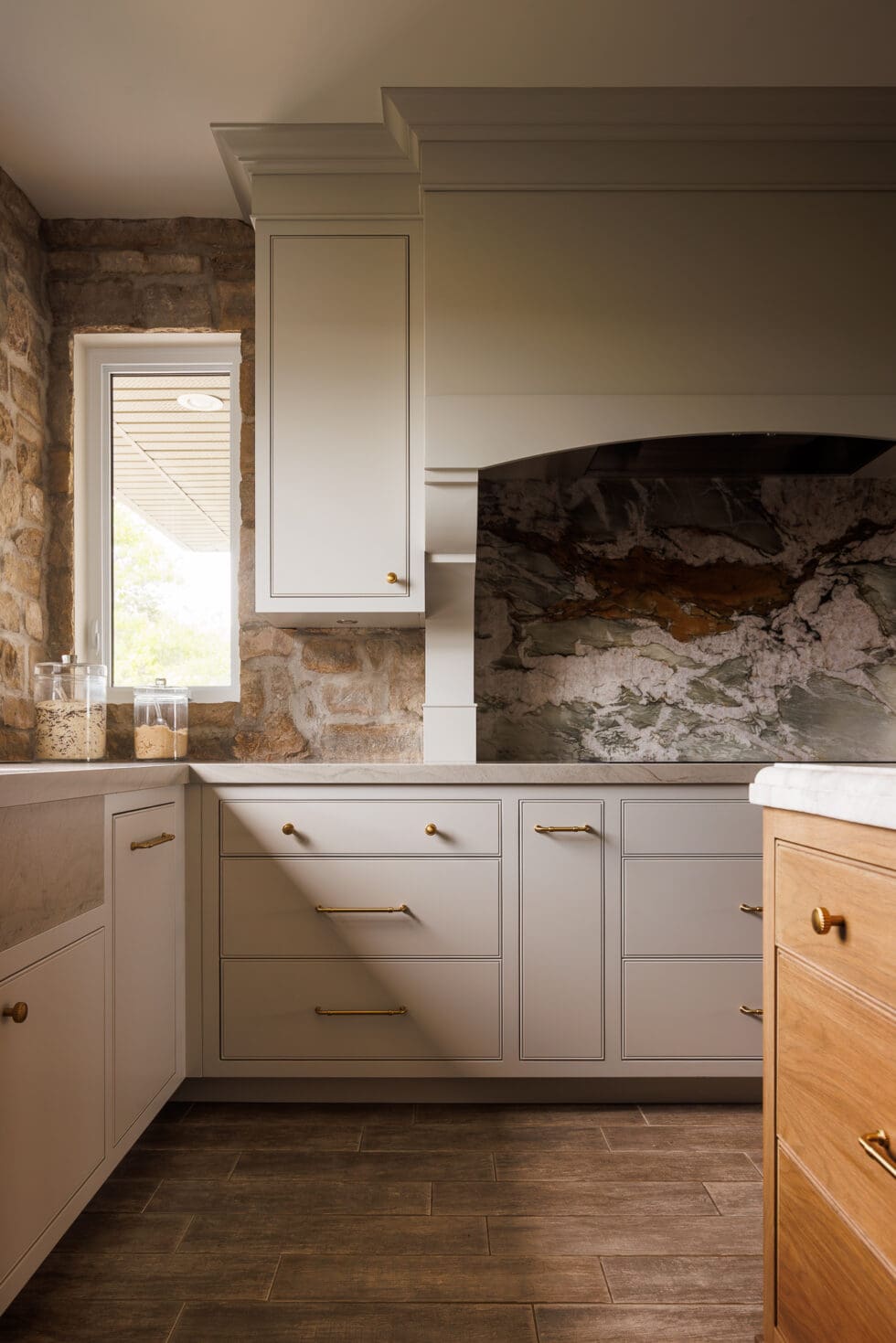 Elegant transitional kitchen featuring light grey cabinets, a dramatic natural stone backsplash, and rustic stone walls, illuminated by a window.