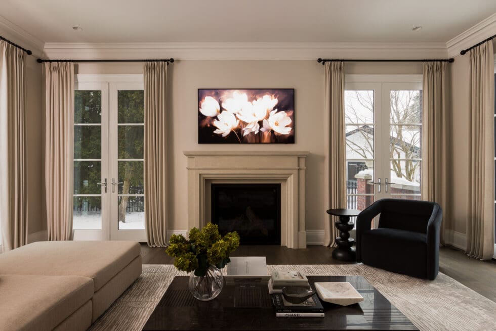 Elegant modern living room with a cream stone fireplace, large TV displaying floral art, beige drapes on French doors, light sectional sofa, and black accent chair.