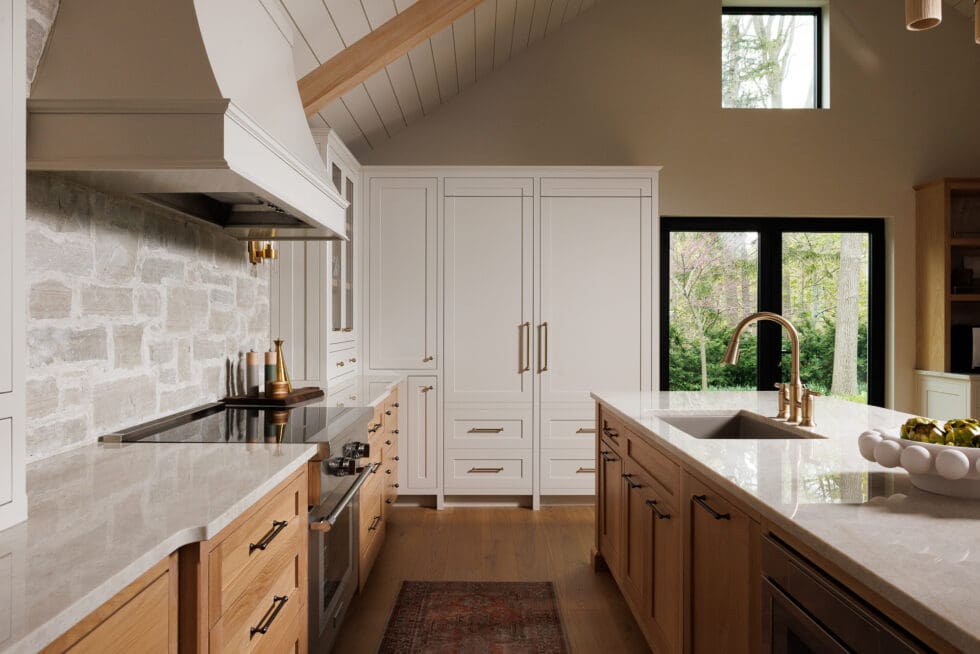 Spacious modern kitchen with light wood lower cabinets, white built-in cabinetry, a stone backsplash, a large island with a brass faucet, and a vaulted ceiling with exposed beams.