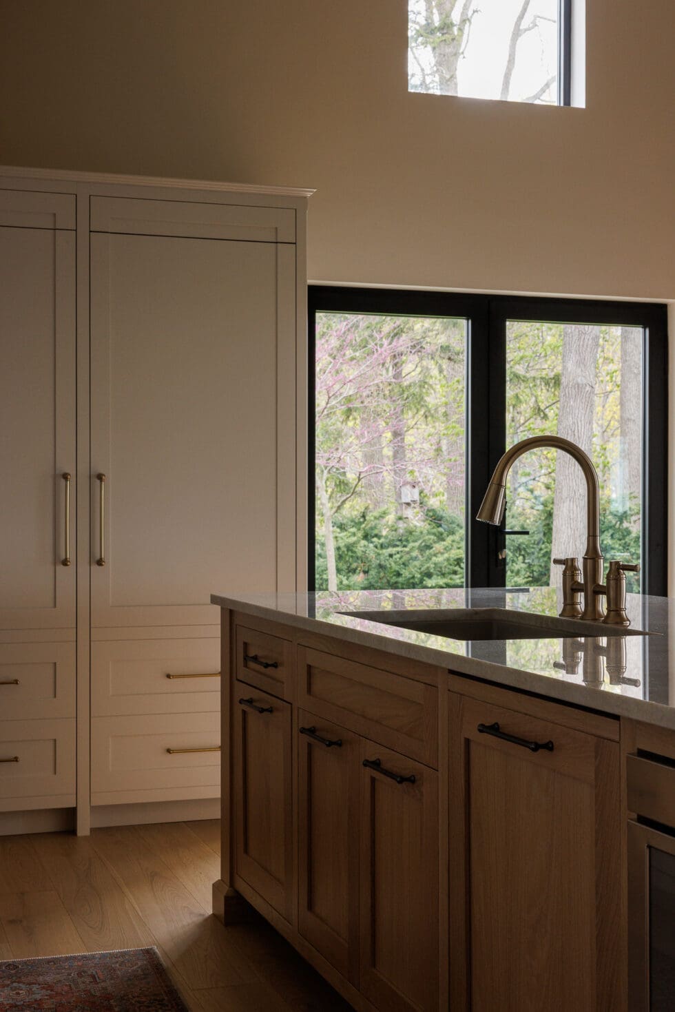 Modern kitchen with large island, brass faucet, built-in cabinets, and expansive windows showcasing a lush outdoor view.