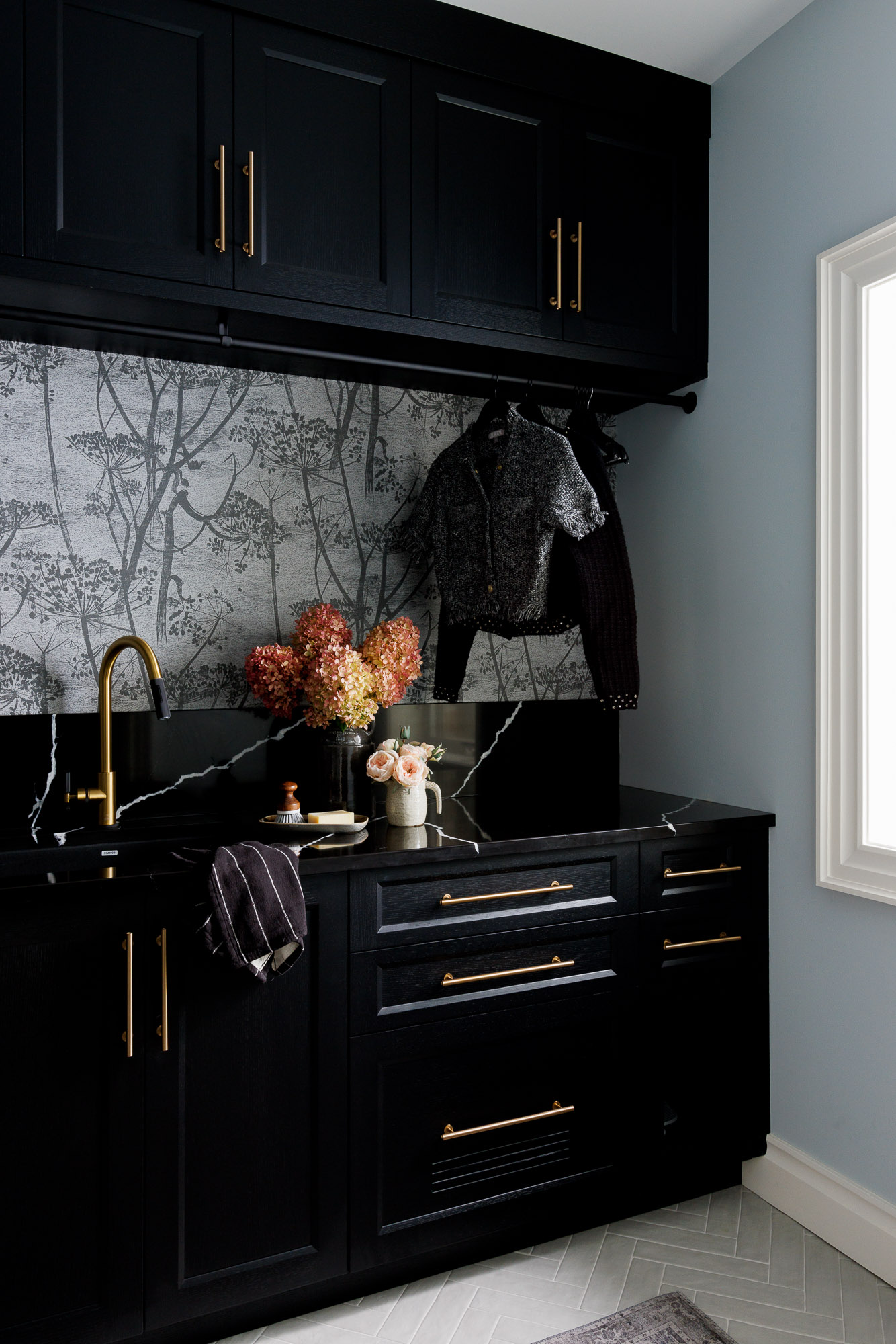 Functional Laundry Storage and Drying Rod A view of the right side of the laundry counter showing black drawers with gold pulls, a black countertop, and clothes hanging on a rod against botanical wallpaper.